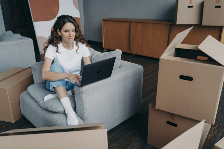 Pensive girl planning house interior design on laptop, sitting with boxes. Moving, flat renovation