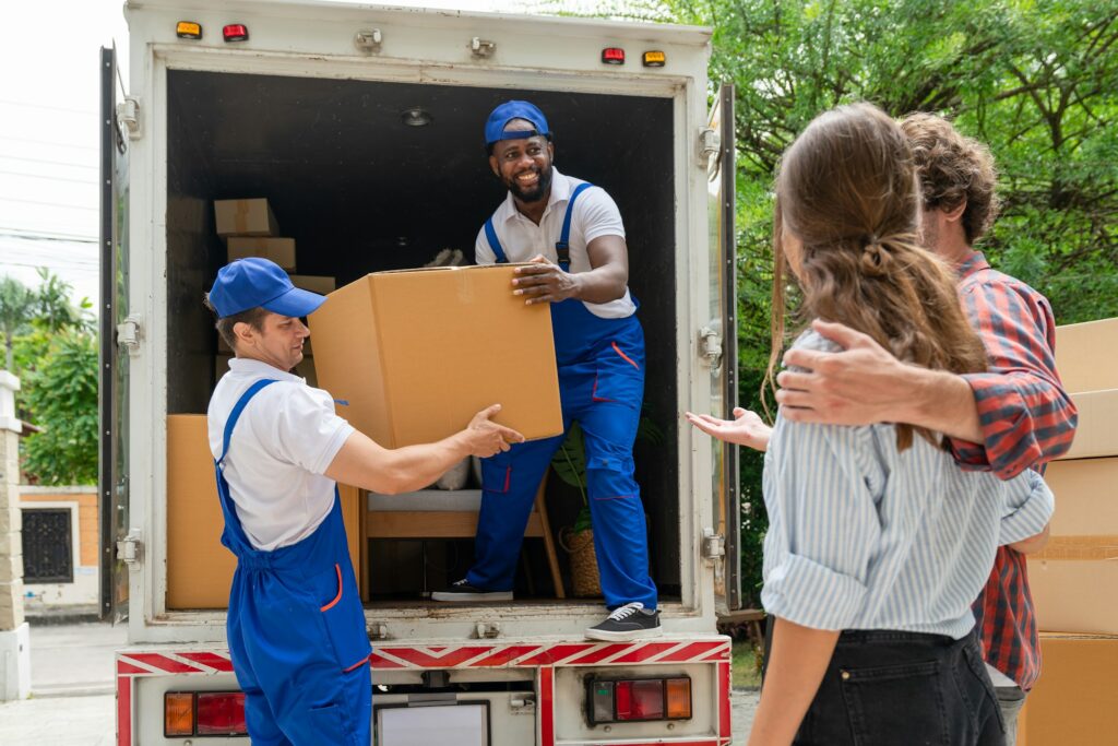 Two men movers in blue uniform lift cardboard box unloading from truck for moving house service